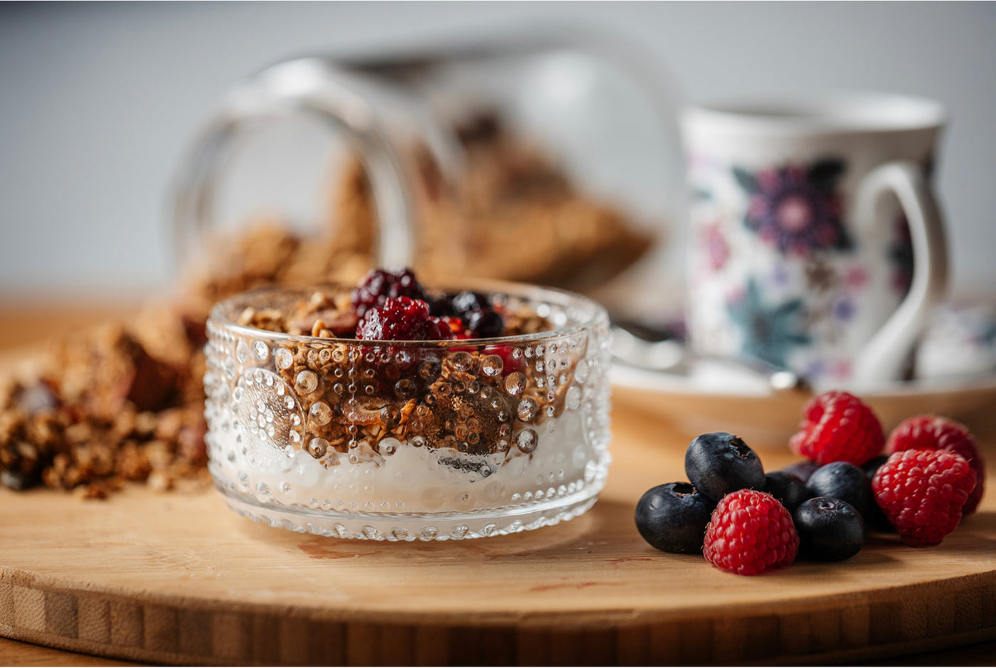 A lifestyle image with a 70s vintage retro bowl containing  Greek yoghourt, Just So Good’s Almond Date granola and a a fruit compote. There are fresh berries in the foreground and a cup of coffee in a retro vintage 70s mug 